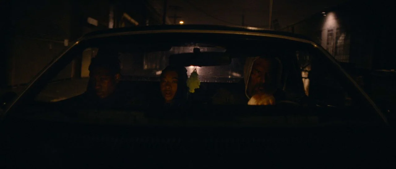 Archenemy (2020) movie still: medium — Three individuals are seated in the front seats of a vehicle at night. The dim environment is lit by outside…