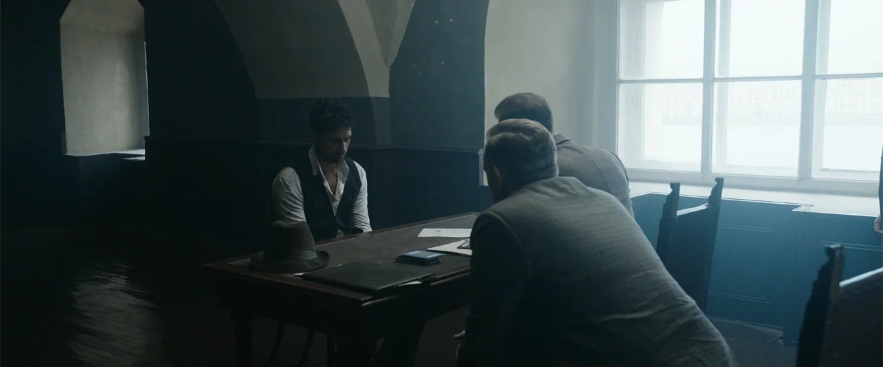 Sardar Udham (2021) movie still: medium — An austere room features stone walls, a wooden table, and a bright window. A man sits at the table while…
