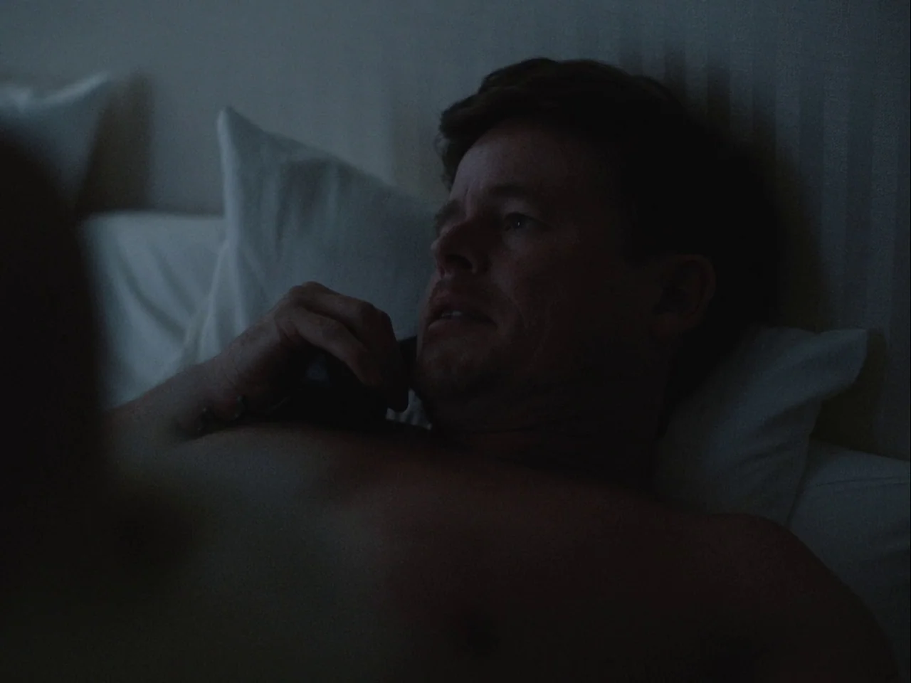 Blonde (2022) movie still: closeup — A dark bedroom with white pillows visible behind a reclining man. A shirtless man lying in bed talks into a…