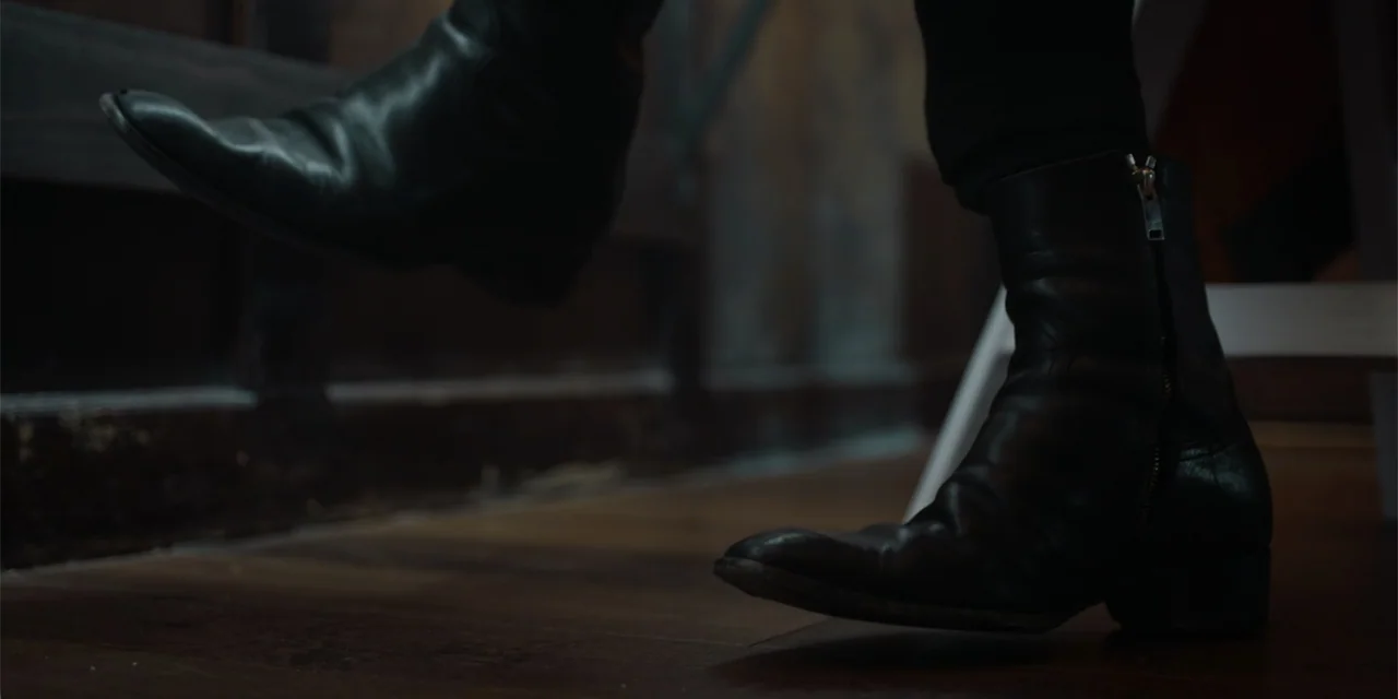 Música (2024) movie still: closeup — A dark, dimly lit room features a worn wooden floorboards with visible dust particles. A pair of black leather…