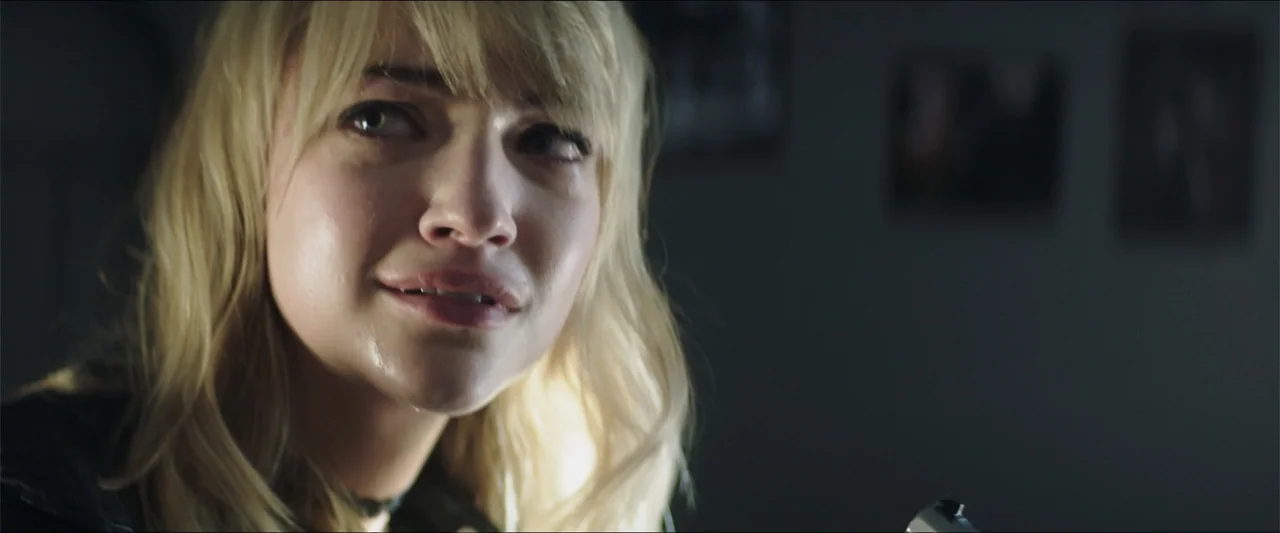 Truth or Dare (2018) movie still: closeup — A dimly lit room with out of focus artwork hanging on a dark wall. A young woman with blonde hair stares…