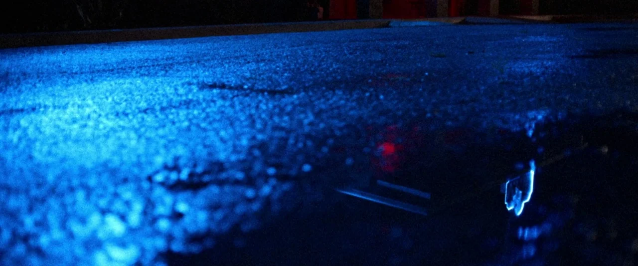 VFW movie still: establishing — A rain-slicked asphalt road at night reflects bright blue and red artificial light sources. The textured surface…