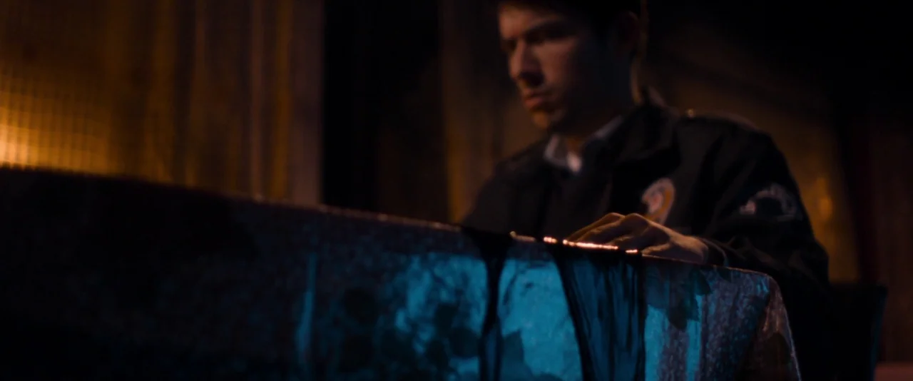 Baskin (2015) movie still: medium — A dimly lit room with industrial textures and a workspace area in low light. A young man in a security uniform…