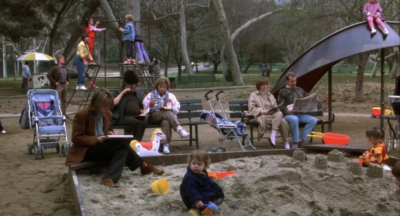 Baby Boom (1987) movie still: wide — A public playground features a large metal slide, a jungle gym, and benches. Multiple adults and children…