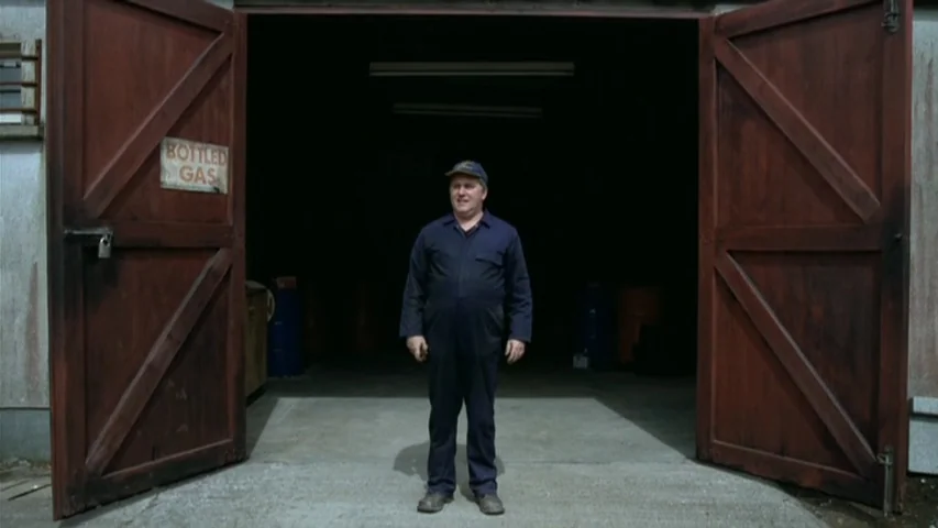 Garage (2007) movie still: fullbody — A man stands before the wide-open double doors of a dark industrial workshop space. He is centered in the…