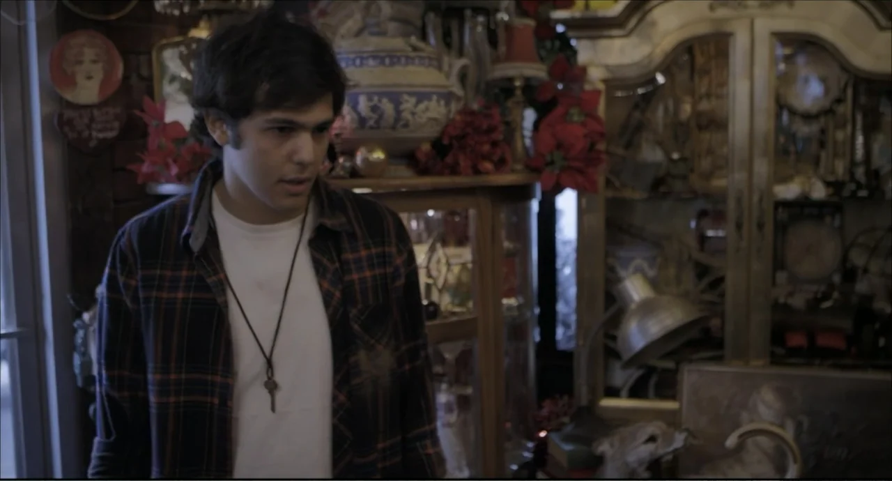 Freedom (2024) movie still: medium — A room interior features a large wood-framed glass cabinet packed with decorative ornaments. A young man stands…