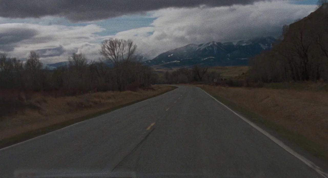 Certain Women (2016) movie still: establishing — An asphalt road extends into a valley flanked by leafless trees and dry grass. Large snow-capped…