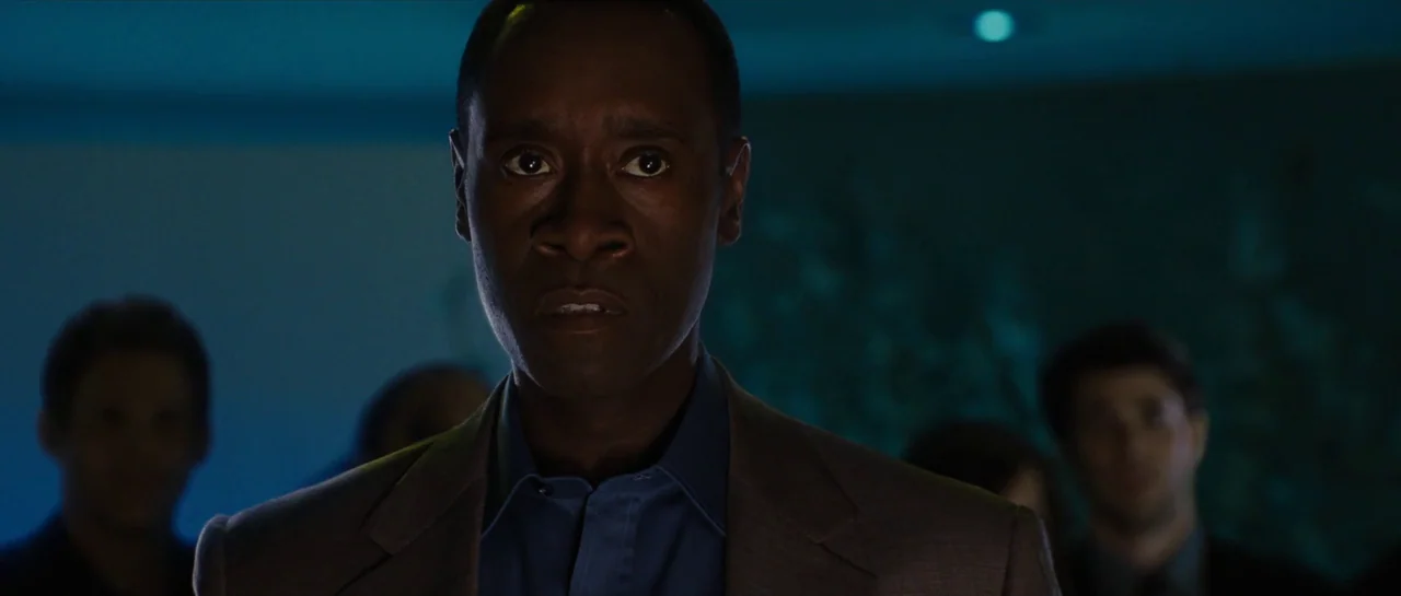 Iron Man 2 (2010) movie still: closeup — A dark, dimly lit room contains several people standing in the background. A man stands in the center…