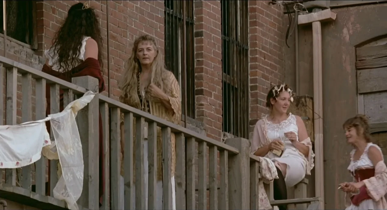 A River Runs Through It (1992) movie still: medium — A group of women in 19th-century attire stands on an outdoor wooden balcony. The women converse…