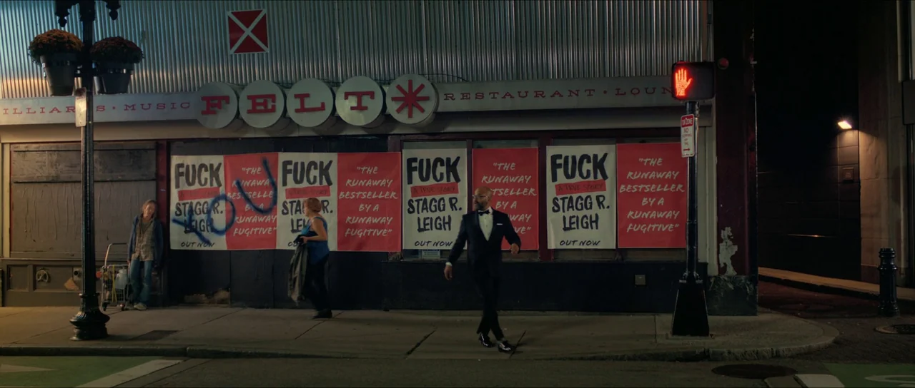 American Fiction (2023) movie still: wide — A city storefront features prominent red advertising posters with bold text and graffiti markings. A man…