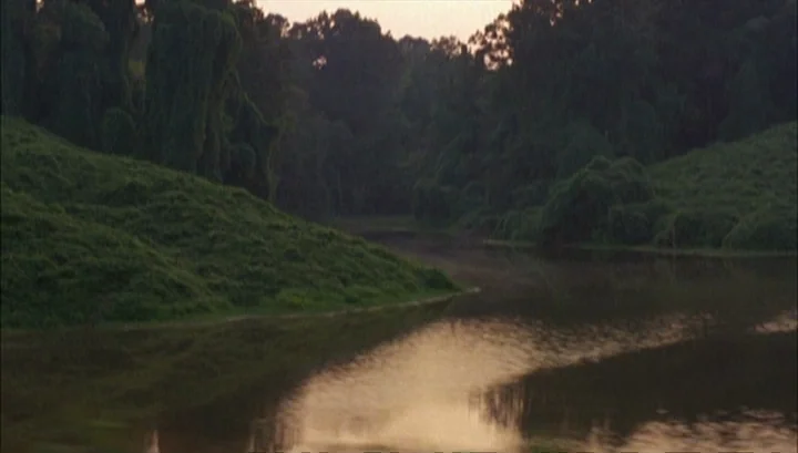 Highway 61 (1991) movie still: establishing — A dark river flows between rolling grassy hills densely covered in thick green vegetation. The sky at…