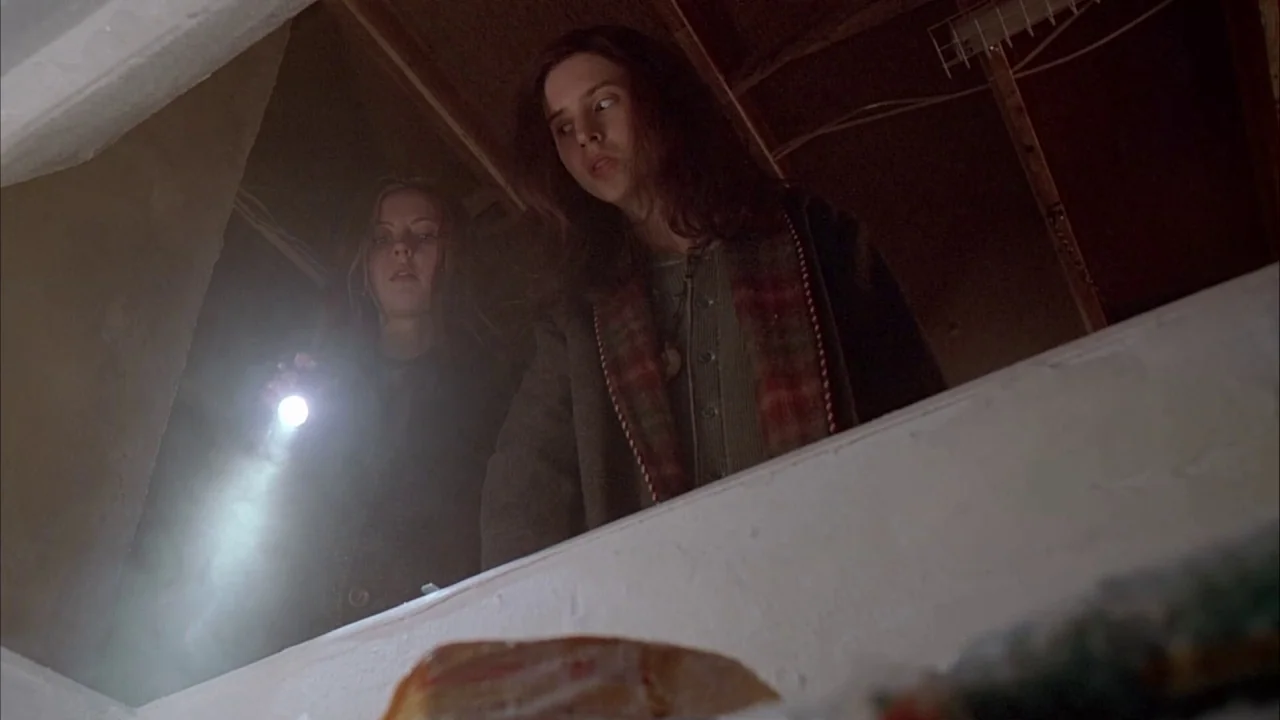 Ginger Snaps (2000) movie still: medium — An unfinished wooden attic space with exposed beams and dim overhead lighting. Two teenage girls stand…