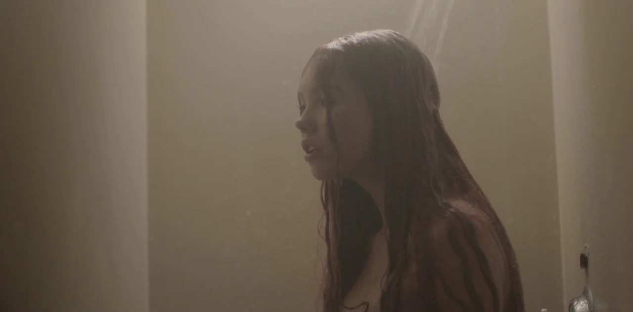 Darlin’ (2019) movie still: closeup — A dimly lit, tiled enclosure with water cascading down from the top right corner. A young woman with long wet…
