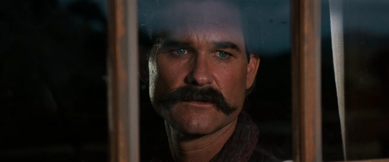 Tombstone (1993) movie still: closeup — A dark, low-key setting with a view through a wooden window frame. A man with a prominent mustache looks…
