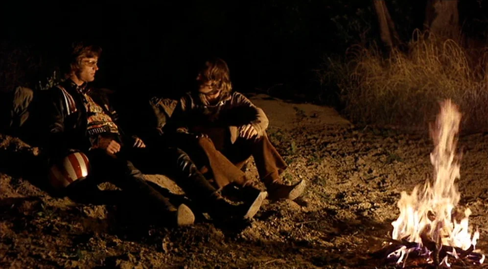 Easy Rider (1969) movie still: medium — Two men sit on the bare, uneven earth at night illuminated only by firelight. They rest near a bright…