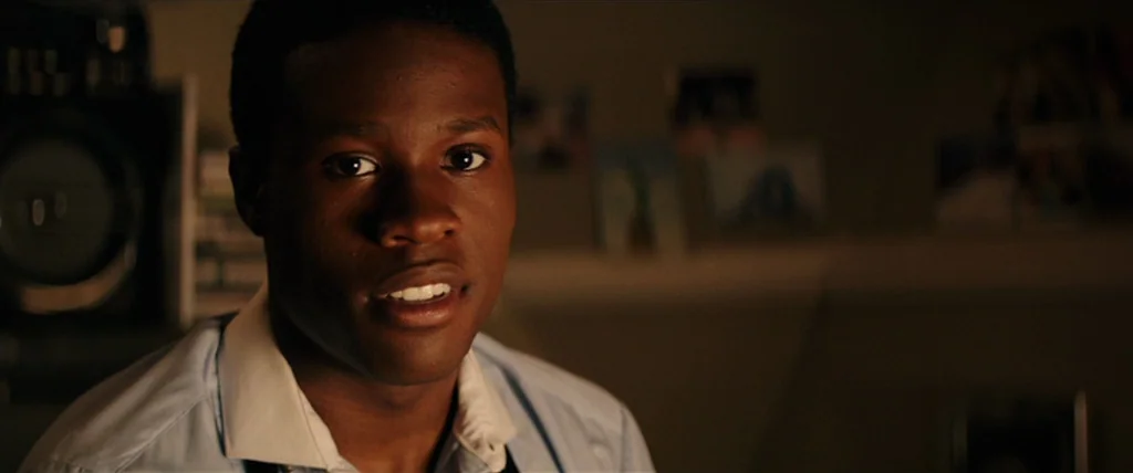 Dope (2015) movie still: closeup — A dimly lit bedroom with out-of-focus posters on the wall in the background. A young Black man looks directly…