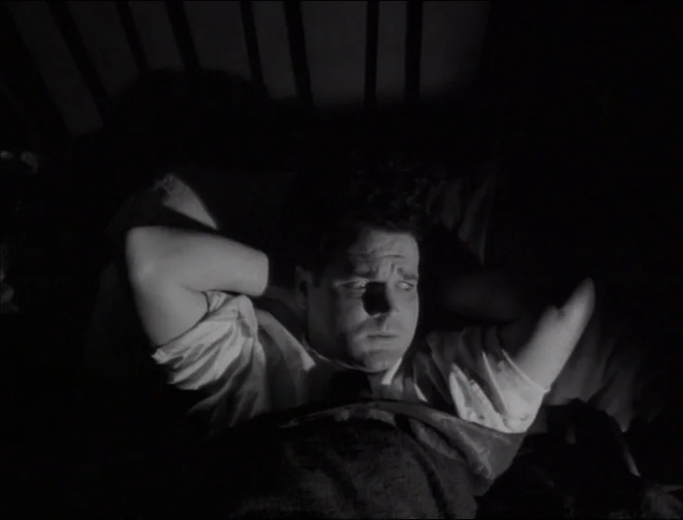 Eraserhead (1977) movie still: medium — A dark bedroom setting with a person lying in a bed. The individual rests with hands clasped behind their…