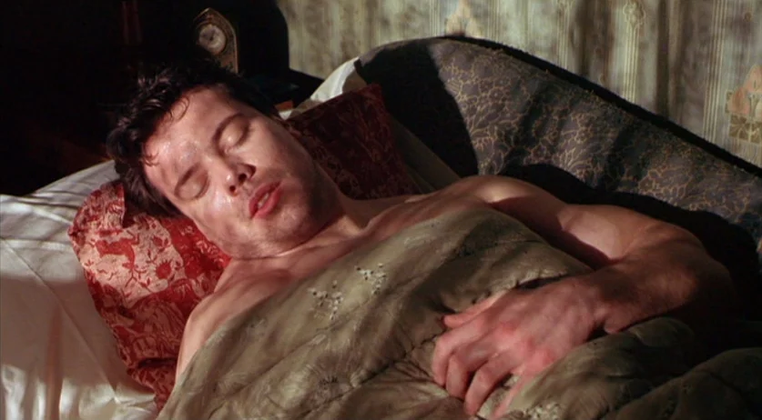 Invincible (2001) movie still: medium — A man sleeps in a bed with dark patterned wallpaper and a vintage clock. He lies under a tan duvet with…