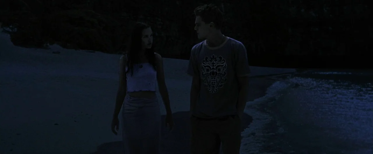 The Beach (2000) movie still: fullbody — A dark, secluded shoreline at night with gentle waves lapping at the sand. A man and a woman walk slowly…