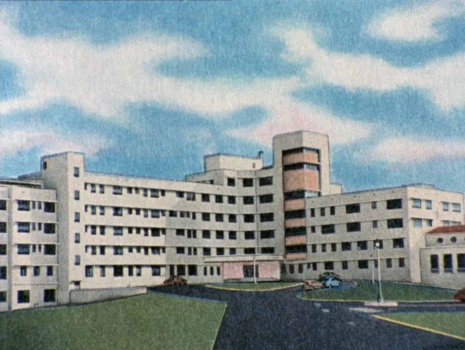 River of Grass (1994) movie still: establishing — A sprawling, modernist-style multi-story hospital building stands under a bright, cloudy blue sky.…