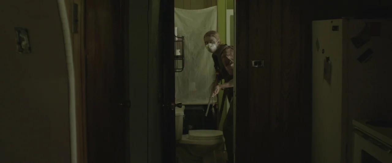 Here Alone movie still: medium — A dimly lit residential hallway with dark wood paneling faces an open door. A man wearing a surgical mask and…