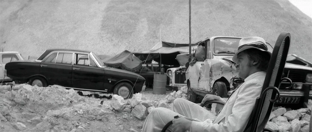 The Last Battle (1983) movie still: medium — A gravel-covered lot contains several damaged cars and a large mound of loose stone. A man in a light…