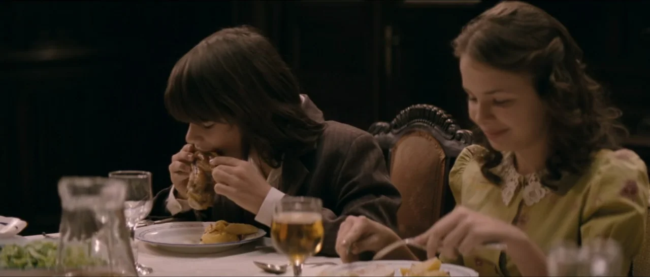 A Farewell to Fools (2013) movie still: medium — A dimly lit room with dark wooden furniture and a set dining table. A teenage boy and girl eat…