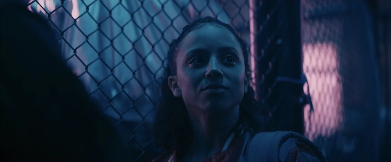 Perfect Addiction (2023) movie still: medium — A woman stands against a chain link fence at night illuminated by neon lights. She looks toward…
