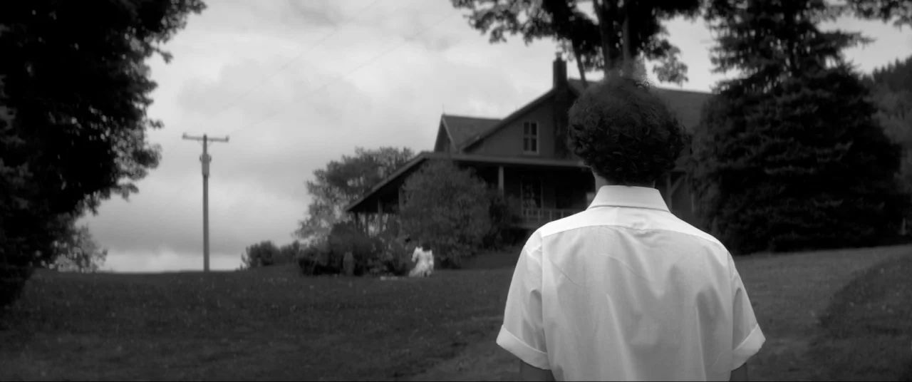 The Eyes of My Mother (2016) movie still: medium — A rural landscape features a two-story house with a porch near dense trees. A man in a white…