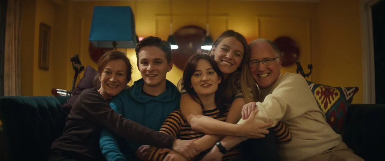 The Rhythm Section (2020) movie still: medium — A family group portrait taken in a yellow-walled room with dark teal furniture. The five people are…
