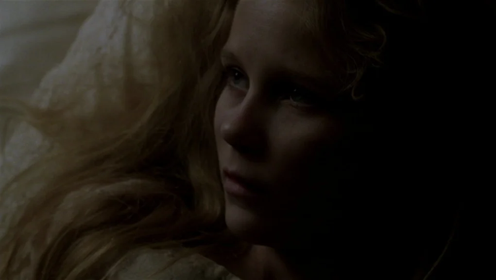 Interview With The Vampire movie still: closeup — A dark, dimly lit bedroom with texture of lace visible on the left side. A young woman with blonde…