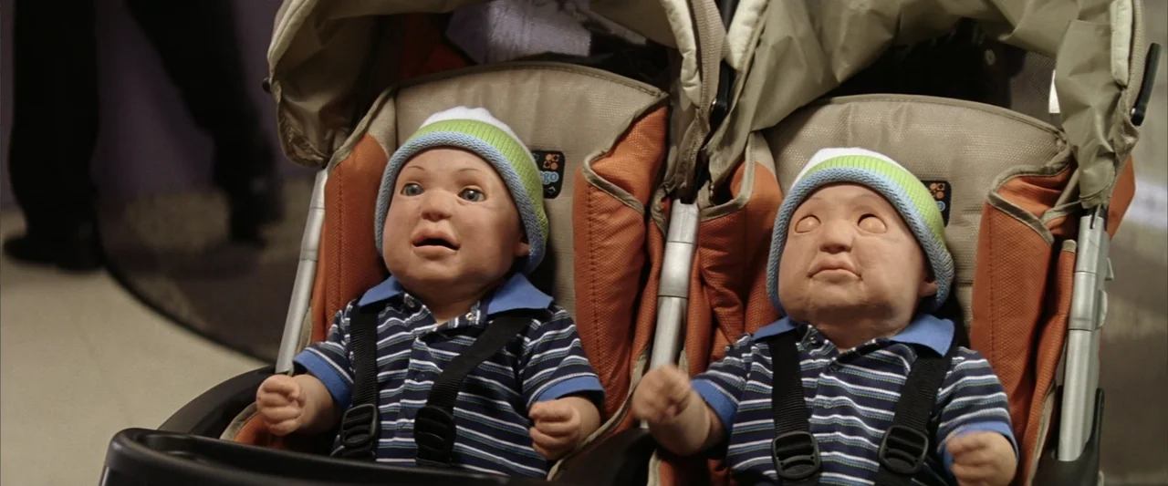 License to Wed (2007) movie still: medium — A double stroller holds two hyper-realistic baby dolls in a brightly lit indoor space. The dolls wear…
