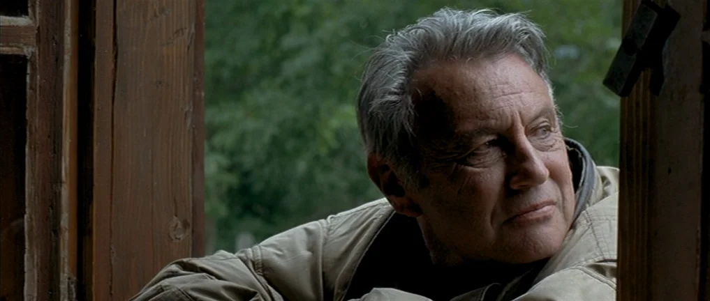 The Intruder (2004) movie still: closeup — An older man looks out from a window frame with weathered wooden molding. Dense green trees are visible…