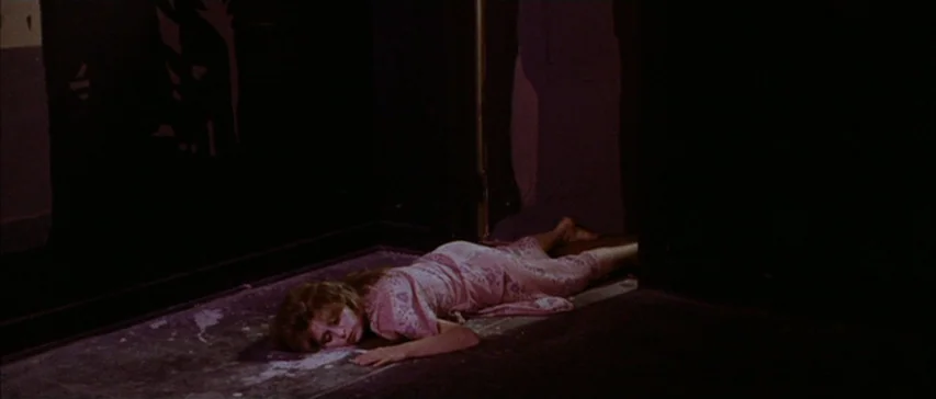 Messiah of Evil (1973) movie still: fullbody — A dimly lit narrow corridor features dark walls and a patterned floor surface. A woman lies flat on…