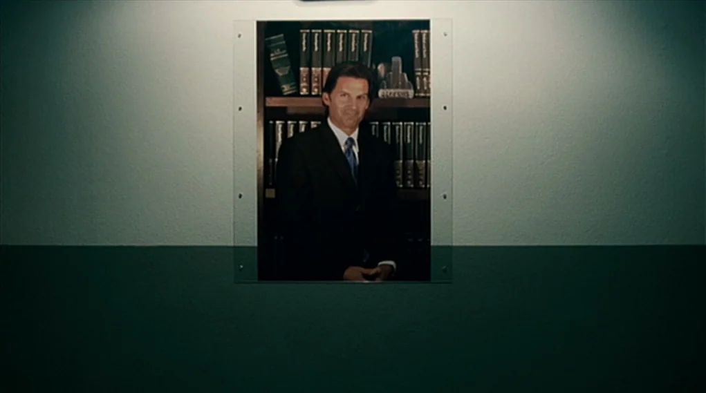 Visioneers (2008) movie still: medium — A two-tone wall features a rectangular portrait mounted behind a clear protective glass sheet. The framed…
