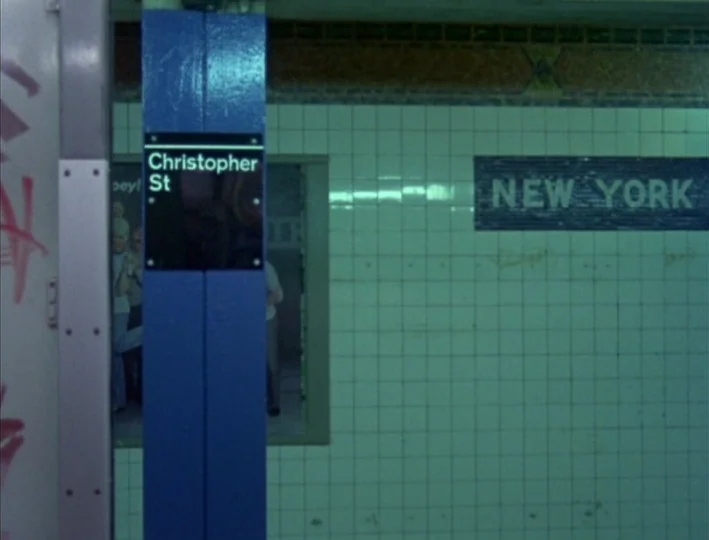 News From Home (1977) movie still: establishing — A subway platform wall features white tiles and a black 'NEW YORK' mosaic sign. A blue vertical…