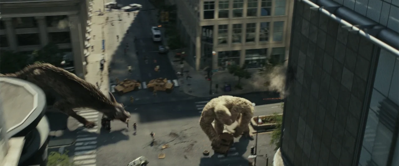 Rampage (2018) movie still: wide — A high-angle view of a city intersection with a giant gorilla and wolf creature. Military vehicles and debris are…