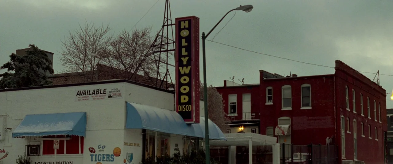 Mississippi Grind (2015) movie still: establishing — A street corner features a white building with an attached Hollywood Disco vertical sign.…
