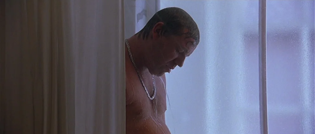 Sexy Beast (2000) movie still: medium — A man stands inside a shower enclosure behind a semi-translucent white plastic curtain. Water droplets cling…