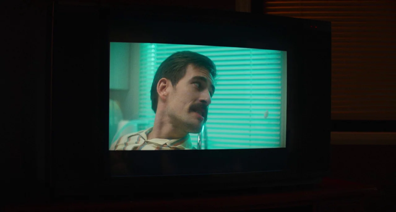 Dual (2022) movie still: closeup — A man appears on a television screen in a darkened room. The screen displays a bright cyan tint showing a man in…