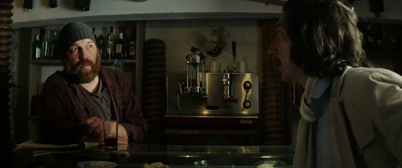 The Man Who Killed Don Quixote (2018) movie still: medium — A dimly lit bar features shelves lined with liquor bottles and a Gaggia machine. Two men…