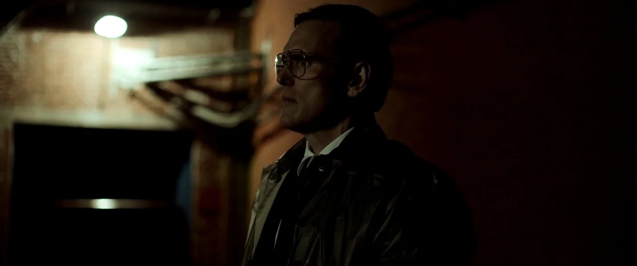 The House That Jack Built (2018) movie still: medium — A dim utility space with industrial piping visible against a rough brick wall. A man in a…
