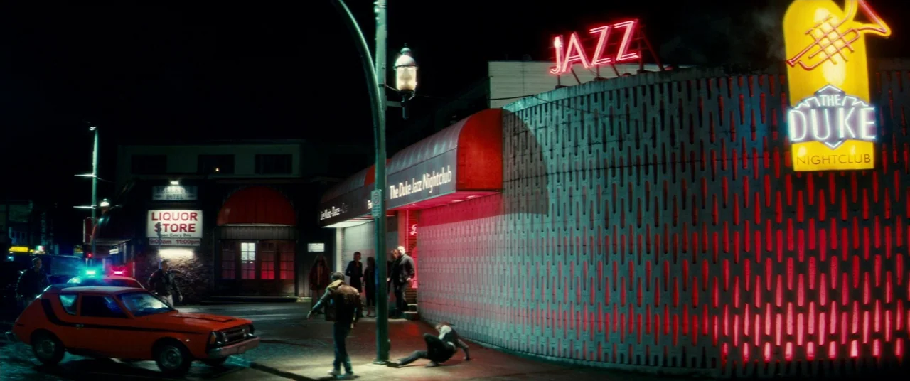 Horns (2013) movie still: wide — A night exterior showing a nightclub facade with a patterned wall and neon signs. A group of people stands on the…