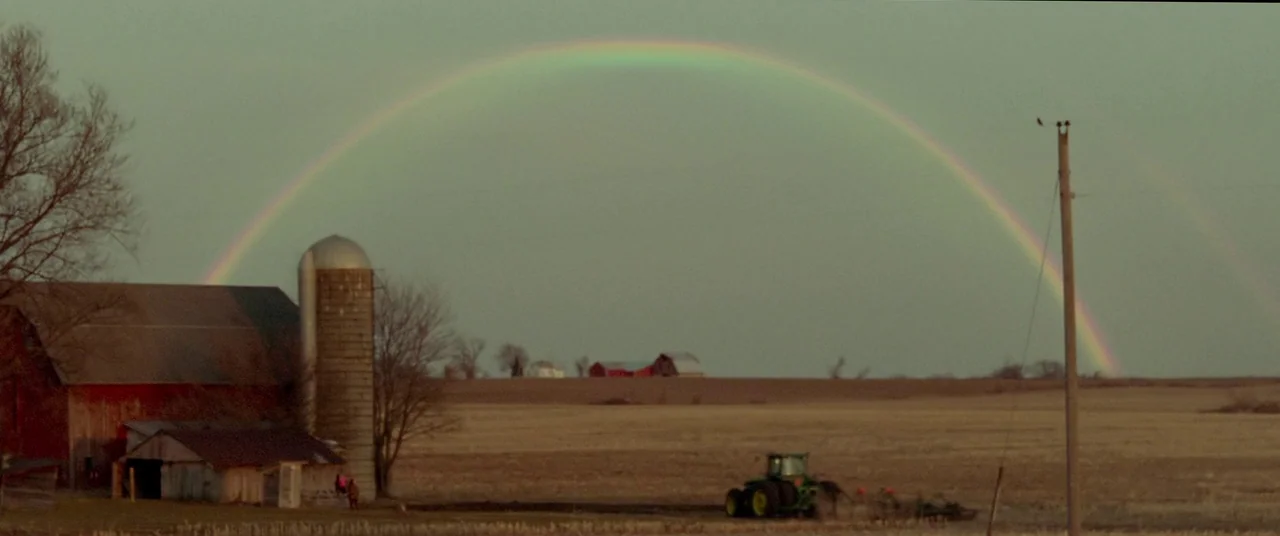 Mississippi Grind (2015) movie still: wide — A rural landscape features a red barn with a metal silo next to fields. A large rainbow spans the…