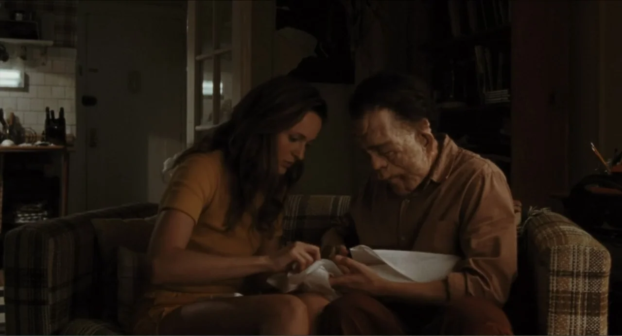 A Different Man (2024) movie still: medium — A dimly lit room with an older man and woman sitting on a sofa. They examine a white piece of cloth…