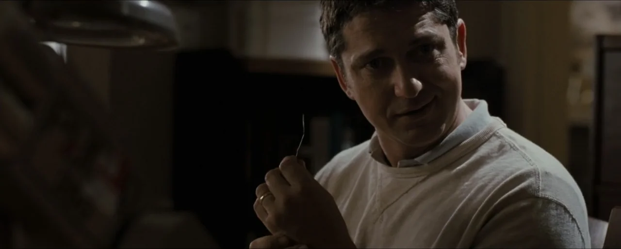 Law Abiding Citizen (2009) movie still: medium — A dark study interior features a man sitting near a desk lamp. The subject looks toward the camera…
