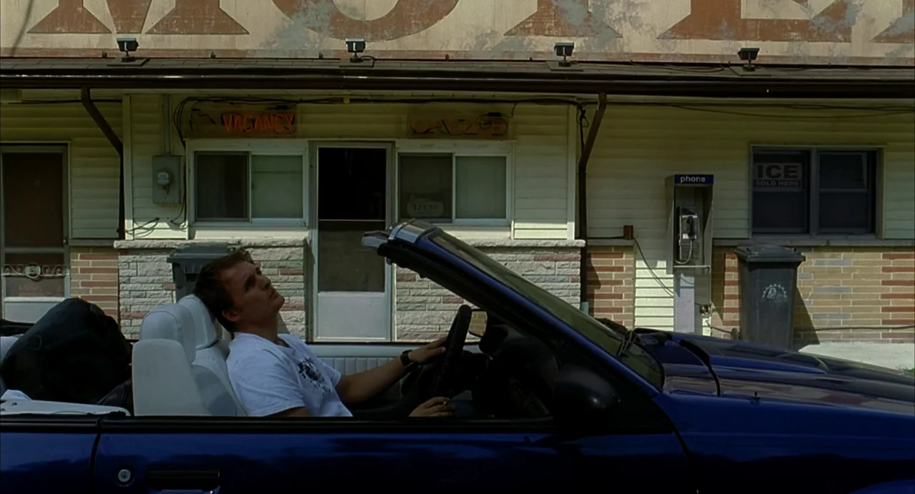 A History of Violence (2005) movie still: wide — A dated roadside motel with peeling painted signage stands behind a blue convertible car. The man…