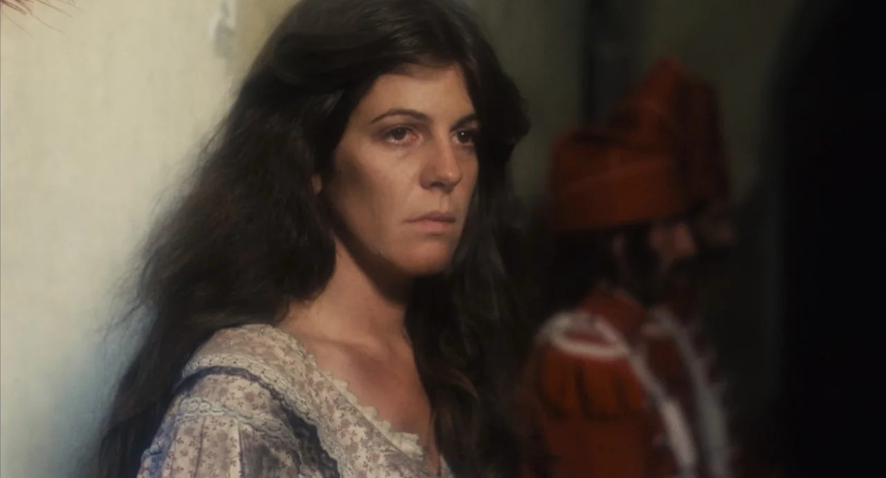 Camila (1984) movie still: closeup — A dimly lit indoor corridor with an off-white wall on the left side. A woman stands in the foreground while a…