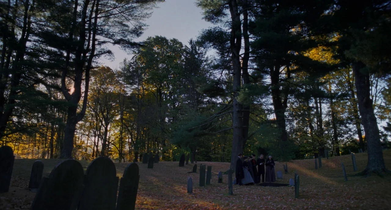 Little Women (2019) movie still: wide — A wooded cemetery with old stone markers scattered among tall autumn trees at sunset. A small group of…