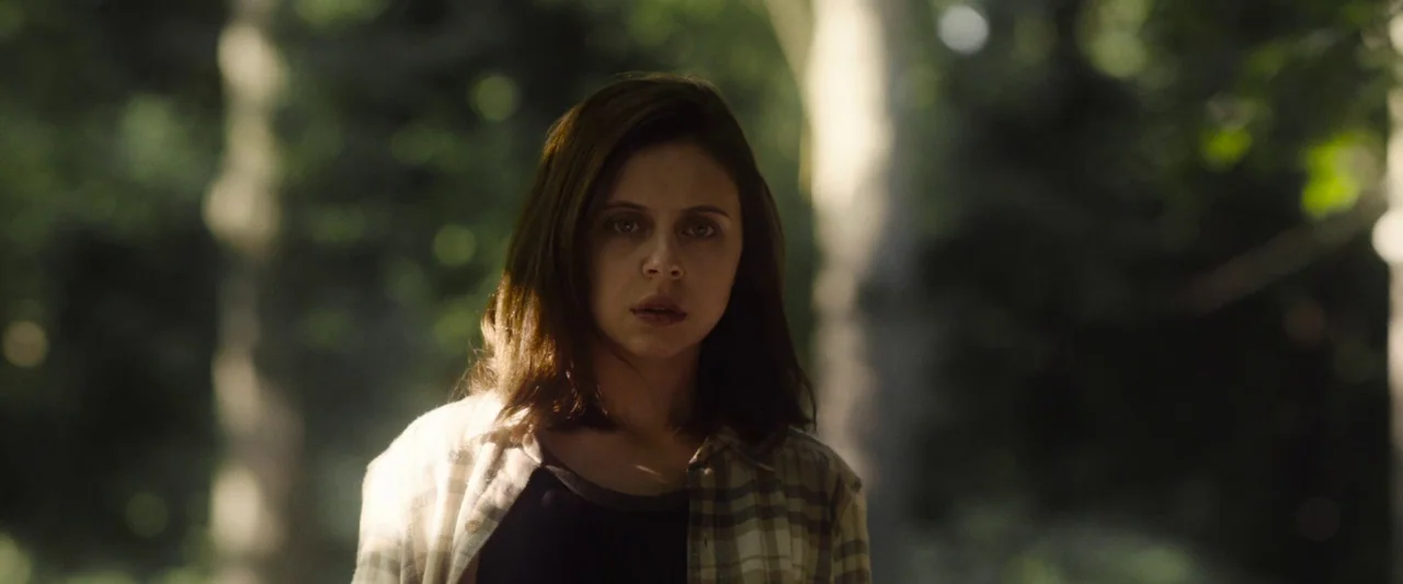 Wildling (2018) movie still: medium — A woman stands in a shaded outdoor location surrounded by tall trees. She looks directly toward the lens in a…